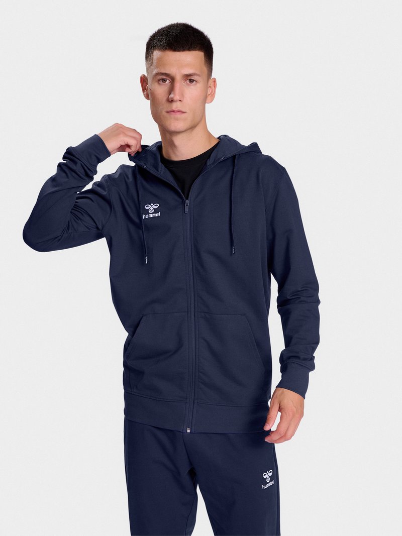 Junger Mann, der die Kapuze seines marineblauen Zip-Up-Hoodies und der passenden Jogginghose mit hummel-Logos auf Brust und Oberschenkel anpasst.