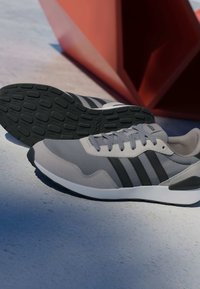 Baskets Adidas grises et noires avec trois bandes noires et semelles intermédiaires blanches, posées sur une surface bleue près d'un objet géométrique orange.