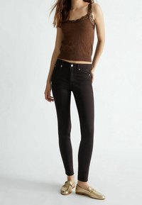 Bruine gebreide tanktop met kantafwerking, gecombineerd met zwarte skinny jeans en gouden loafers, met een soepele textuur en een getailleerd ontwerp.