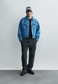 Geacă de blugi albastru deschis cu două buzunare pe piept, asortată cu pantaloni negri cu accente pe cusăturile laterale și adidași negri. Căciulă albă pe cap.