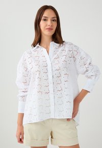 White embroidered blouse with a button-up front, featuring eyelet cutouts and a relaxed fit. Long sleeves and a curved hem.