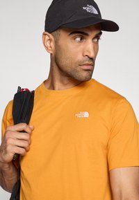 Πορτοκαλί βαμβακερό μπλουζάκι με λευκό κεντημένο λογότυπο στο αριστερό στήθος. Συνδυάζεται με μαύρο καπέλο και μαύρο ιμάντα σακιδίου.