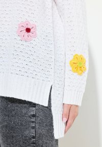 Maglione bianco a maglia con trama larga, con applicazioni di fiori rosa e gialli sulle maniche. Orlo a maglia con spacchi laterali per maggiore comfort.