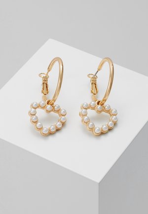 Guldcreoler med hængende hjerteformede charms dekoreret med små hvide perler, vist på en hvid geometrisk overflade.