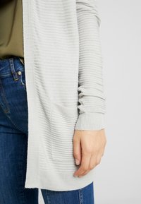 Světle šedý pletený cardigan s žebrovanou texturou, s otevřeným předním dílem a kapsou; nošený přes olivové tričko a kombinovaný s modrými džíny.