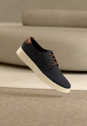 Baskets décontractées noires avec lacets marron et languette au talon, semelle en caoutchouc crème, présentées flottant au-dessus d'un tapis beige devant un canapé.