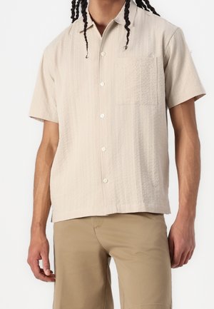 Man draagt een lichtbeige, kortärmelig hemd met borstzak en beige broek, staand tegen een effen witte achtergrond.