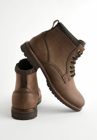 Brown leather ankle boots with black laces, low heel, and textured wool lining. Features gold eyelets and contrasting stitching.