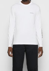 Mann trägt ein weißes Langarmshirt von Calvin Klein mit Logo auf der Brust und Text auf dem linken Ärmel, kombiniert mit schwarzen Hosen.