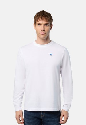 Uomo con maglietta bianca basic a maniche lunghe con piccolo logo circolare blu sul petto, abbinata a jeans blu scuro, in piedi su uno sfondo chiaro.