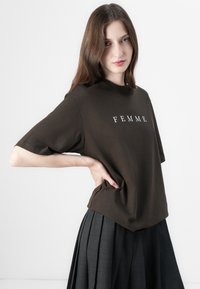 Donkerbruine katoenen t-shirt met de tekst "FEMME" in het wit, met een relaxed fit en korte mouwen, gecombineerd met een geplooide donkere rok.