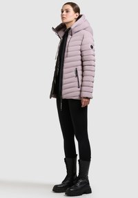 Lys lilla pufferjakke med hætte, vandret kviltet design og sort hardware. Båret over en sort top, parret med sorte leggings og støvler.