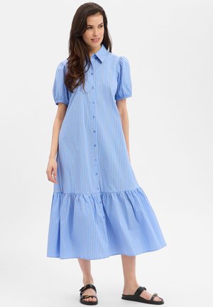 Kobieta ubrana w jasnoniebieską, paskowaną sukienkę midi na guziki z bufkami, stojąca i uśmiechająca się, w połączeniu z czarnymi płaskimi sandałami.