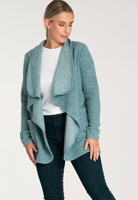 Widok z przodu na turkusowy kardigan z otwartym przodem, teksturowana tkanina, długie rękawy i warstwowy design, noszony na białej bluzce.