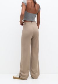 Pantalon beige à jambes larges avec une texture lisse, doté d'une taille haute, de poches latérales et d'une coupe décontractée. Associé à des baskets de couleur claire.