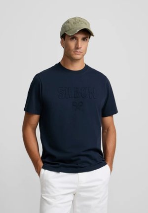 Hombre con una camiseta azul marino oscuro con texto en relieve "SILBON" y raquetas de tenis cruzadas, pantalones blancos y una gorra de béisbol verde clara.