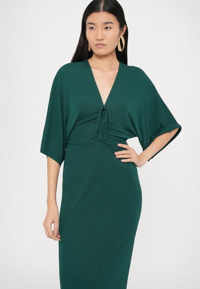Πράσινο φόρεμα από απαλό ύφασμα με βαθύ V λαιμόκοψη και ζώνη δέσιμο στη μέση. Διαθέτει φαρδιά μανίκια και εφαρμοστή σιλουέτα μέχρι το πάτωμα.