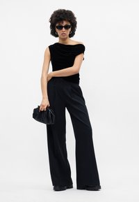 Μαύρη βελούδινη τοπ με σχέδιο off-shoulder, συνδυασμένο με ψηλόμεσες μαύρες παντελόνες φαρδιές και μια μικρή μαύρη τσάντα συλλογής.