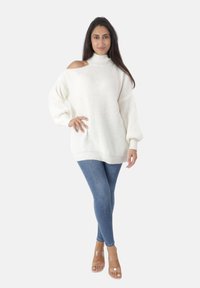 Femme debout portant un pull en maille épaisse blanc avec une épaule dégagée, un jean skinny bleu et des sandales à talons avec des bretelles transparentes.