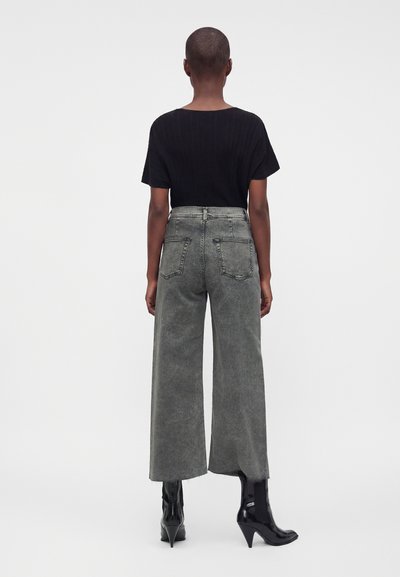 Personne aux cheveux courts portant une chemise noire à manches courtes, un jean gris taille haute à jambes larges, et des bottines noires à talons, debout, vue de dos.