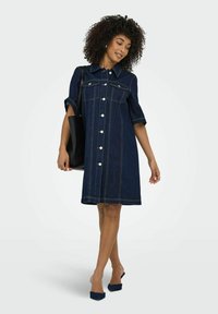 Denim skjortklänning i mörkblått, med korta ärmar, knappstängning och två bröstfickor. Slät textur med kontrasterande gul sömnad.