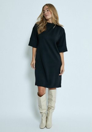 Black knee-length dress with a high neck, relaxed fit, and short sleeves. Paired with cream knee-high boots with a slouchy design.