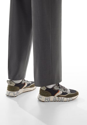 Gambe inferiori che indossano pantaloni larghi grigi e sneakers con suole bianche, caratterizzate da grande testo "BLANCHE" su di esse, in piedi su una superficie bianca.