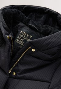 Veste matelassée bleu marine foncé avec un intérieur en peluche noire et des accessoires dorés, dotée d'une fermeture éclair et d'une étiquette portant la mention "NEXT".