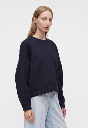 Ženska z ravnimi blond lasmi nosi ohlapno črno majico s kapuco in svetlo modre jeans hlače, stoji pred enobarvnim belim ozadjem.