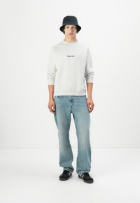 Grijze sweatshirt met ronde hals en zwarte tekst, lichtblauwe losvallende jeans, zwarte bucket hat en zwarte sneakers met witte accenten.