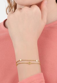 Bracciale oro con design a doppia catena; presenta piccoli accenti a forma di stella e di stella ritagliata. Indossato su una mano contro uno sfondo rosa.