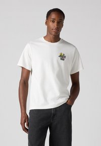 Hvit bomulls t-skjorte med et fargerikt grafisk design på venstre bryst, sammen med mørkegrå denimjeans. Enkel rund hals og korte ermer.