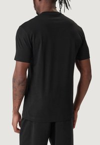 T-shirt nera a maniche corte in cotone; presenta uno scollo rotondo e un orlo dritto. È visibile la schiena del modello, con un leggero dettaglio sulla spalla.