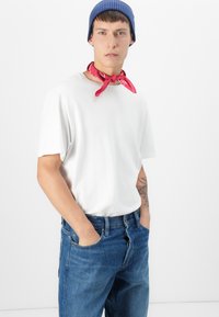 Camiseta blanca de manga corta, gorro de punto azul y bufanda roja al cuello. Jeans de mezclilla azul con bolsillos visibles y un tatuaje en la muñeca. Fondo liso.