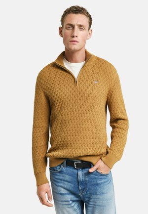 Senapsgul stickad tröja med ett texturerat mönster, dragkedja vid halsen och ribbade ärmar, parat med ljusblå jeans och ett svart bälte.