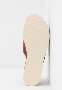 Chaussure en daim de couleur brun rougeâtre avec une semelle en caoutchouc crème texturée présentant des rainures horizontales et un bout arrondi. Marquage de la taille visible.