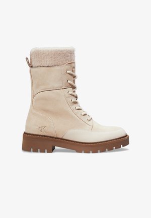 Beige ruskind ankelstøvler med snørebånd, blød pelskrave, gummisål og lyse farver. Har et logo på ydersiden.
