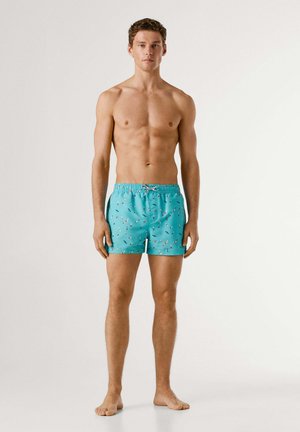 Mees seisab paljajalu, kandes türkiissiniseid ujumispükse, millel on väikesed mitmevärvilised linnupildid, tavalise valge tausta ees.