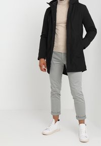 Svart vattentät jacka med huva, framdragkedja och sidofickor, tillsammans med ljusgrå uppvikta byxor och vita sneakers.