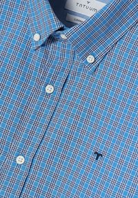 Camicia a quadri blu e bianca con colletto button-down, dotata di due bottoni bianchi e un piccolo logo scuro sul petto. Design a maniche corte.