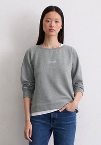 Mujer joven con cabello largo y negro lleva un sudadera gris de Marc O’Polo y jeans azules, de pie contra una pared blanca simple.