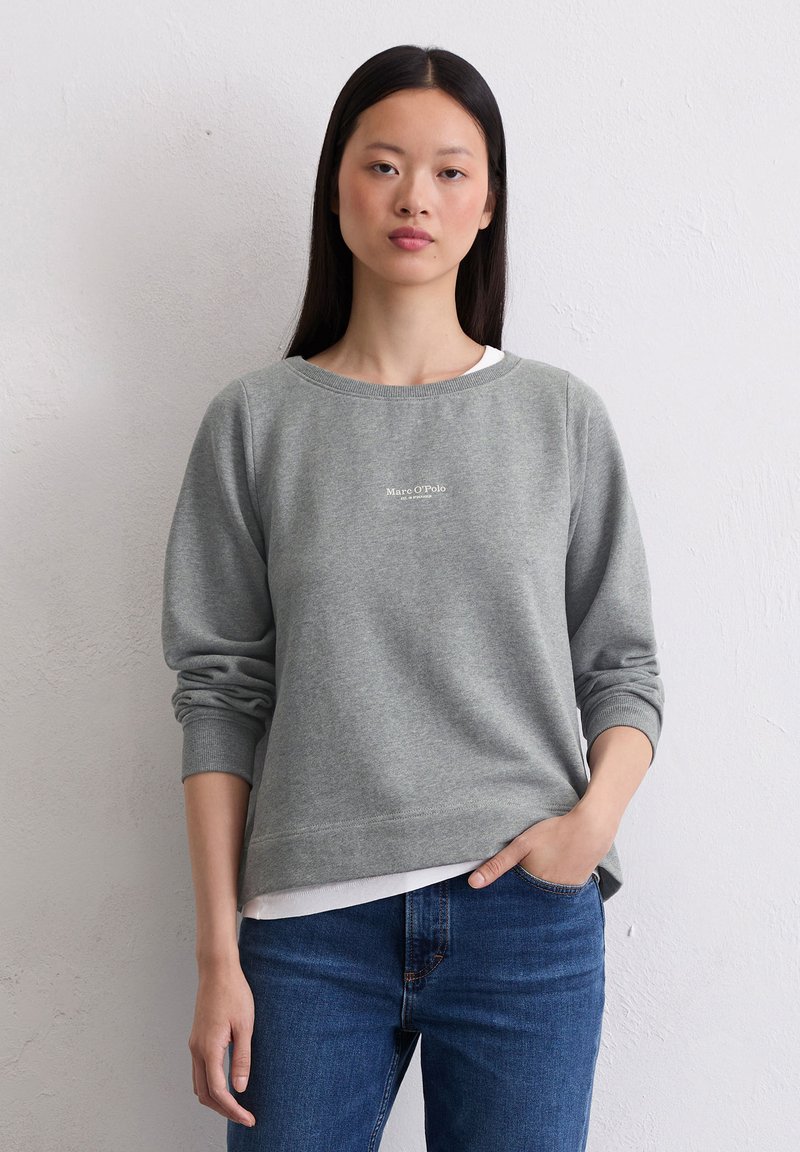 Mujer joven con cabello largo y negro lleva un sudadera gris de Marc O’Polo y jeans azules, de pie contra una pared blanca simple.