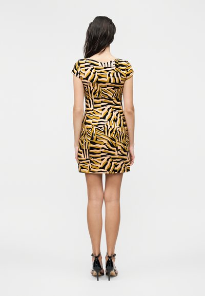 Γυναίκα που στέκεται με την πλάτη, φοράει ένα κοντομάνικο φόρεμα μέχρι το γόνατο με κίτρινο, μαύρο και μπεζ αφηρημένο μοτίβο και μαύρα σανδάλια με ψηλό τακούνι.