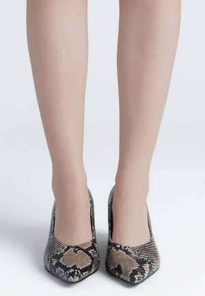 Piernas con zapatos de tacón alto de punta afilada con un patrón de piel de serpiente marrón y negro sobre un fondo blanco.