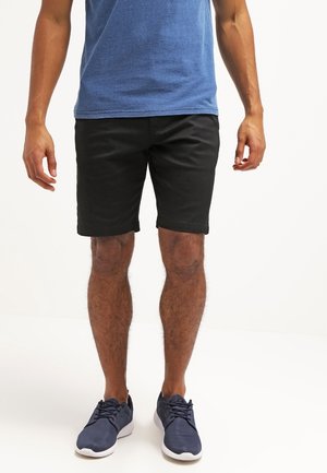 Mann trägt schwarze Shorts, blaues T-Shirt und marineblaue Turnschuhe, steht vor einem schlichten weißen Hintergrund.