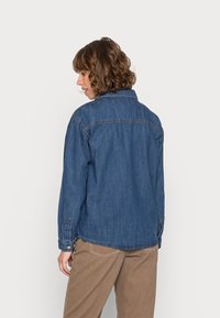 Camisa de ganga com colarinho clássico e punhos com botão, em azul escuro. Apresenta um corte reto com detalhes de costura visíveis e um yoke nas costas.