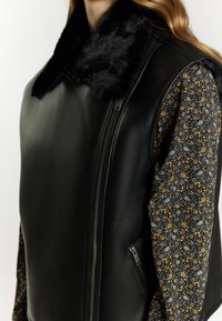 Zwarte leren vest met een zachte zwarte kraag, voorzien van een zijrits en mouwdetails met een bloemenpatroon in wit, geel en groen.