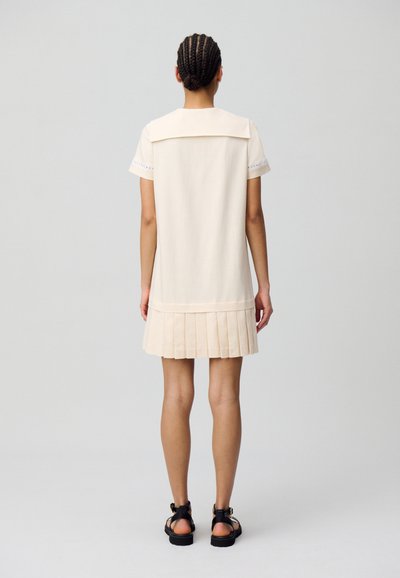 Vestido corto color crema con dobladillo plisado, presenta un cuello amplio y mangas cortas con detalles decorativos. Lleva sandalias negras.