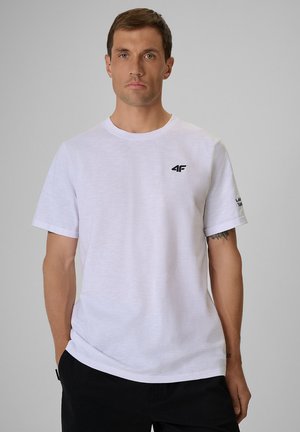 Mężczyzna ubrany w zwykły biały t-shirt z krótkim rękawem i małymi czarnymi logo na piersi i lewym rękawie, stojący na jednolitym szarym tle.