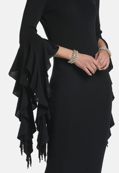 Vestido negro ajustado con dramáticas mangas de volantes en capas, mostrado sobre torso y brazos que llevan dos pulseras doradas con joyas.